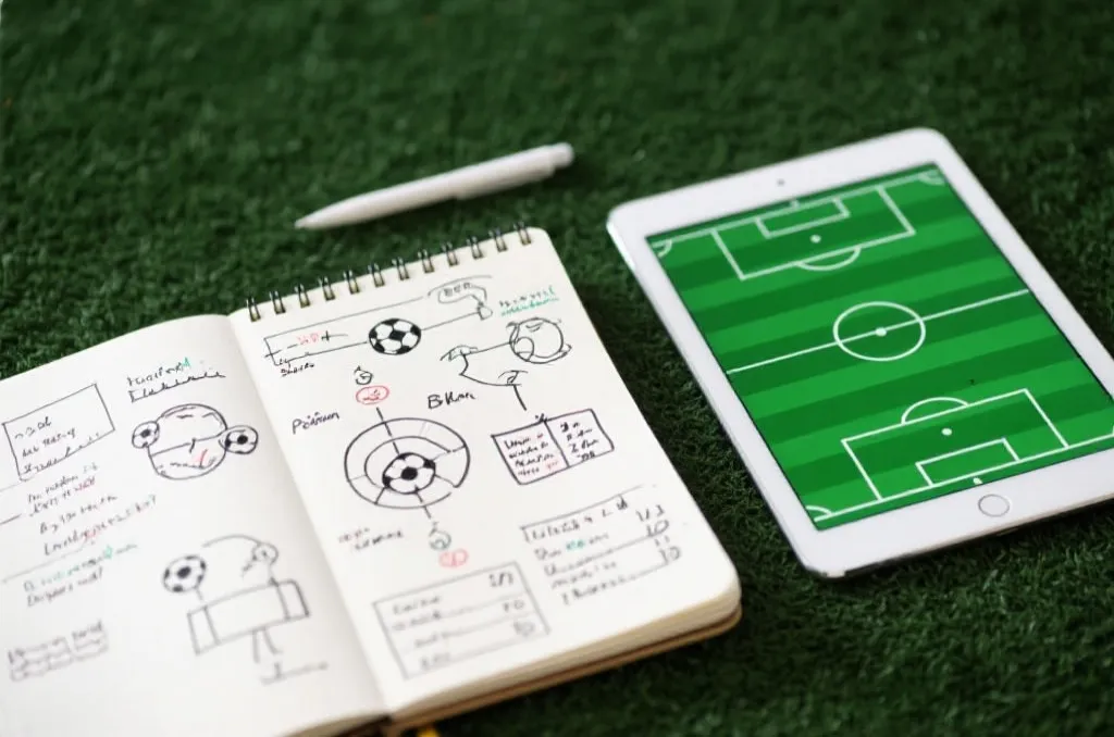 An open notebook with hand-drawn soccer tactical diagrams and statistical notes next to a tablet showing a generic green field layout